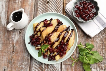 Tasty crepes with cherries, chocolate and mint served on wooden table, flat lay