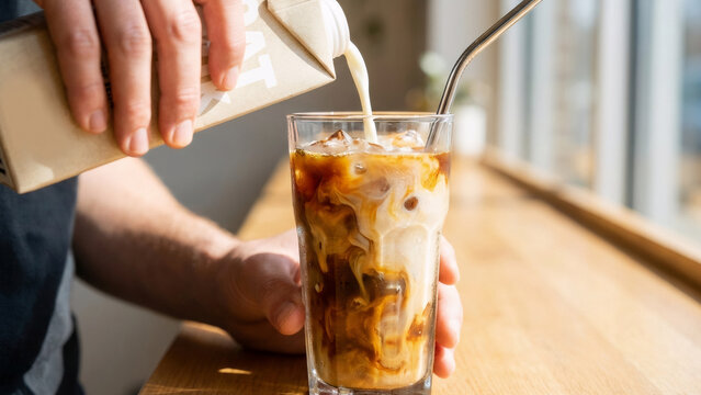 Man pouring oat milk into iced coffee with a metal straw in a glass. Refreshing cold drink for summer. Plant based beverage concept.