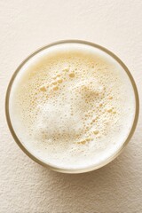 Vista dall'alto ravvicinata di un bicchiere rotondo trasparente pieno di latte bianco e spumoso, appoggiato su una superficie beige 