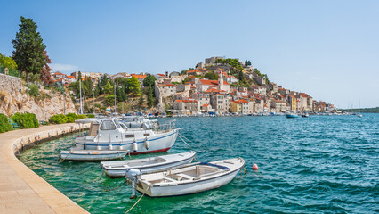 The coastal town of Sibenik in Croatia sits on the seafront, with hilltop houses and St. Michael's Castle. Boats moored along the shore, viewed from the walking path on a sunny summer day.