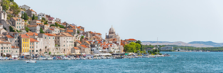 Fototapeta premium The coastal town of Sibenik in Croatia, nestled on the seafront with hilltop houses and the Cathedral of St. James, is seen from a walking path on a sunny summer day.