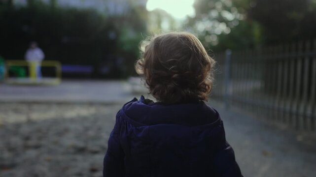 Toddler standing in park looking toward playground with soft curls catching light creating gentle outdoor moment showing curiosity exploration and natural childhood innocence on cool afternoon