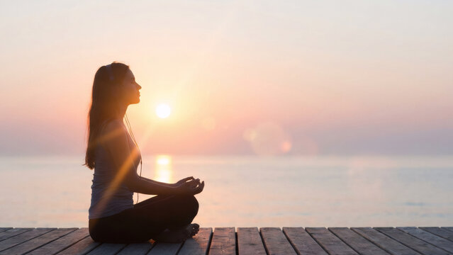 Woman meditating on wooden pier at sunset with headphones. Serene spiritual practice and mindful relaxation by the tranquil ocean.