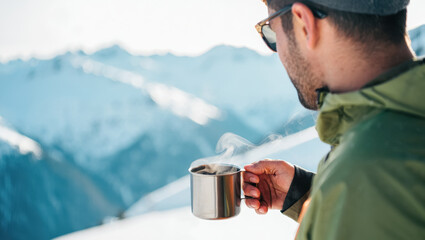 A man in a beanie and sunglasses holding a steaming metal cup while overlooking a soft-focused snowy mountain landscape, with warm sunlight highlighting the vapor and winter textures