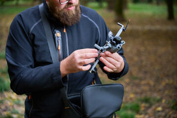 A man with a beard inspects a drone in a green forest. Sunlight filters through the trees as he prepares for an exciting aerial adventure.