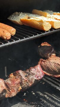 Thick beef steaks sear on a hot grill while bread toasts on the upper rack. Smoke, fat, and char mark a delicious outdoor barbecue scene.