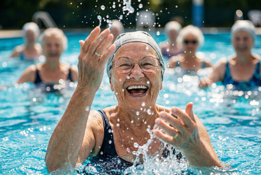 Joyful senior woman with glasses splashing water and laughing during a fun aqua aerobics class with friends in the swimming pool.