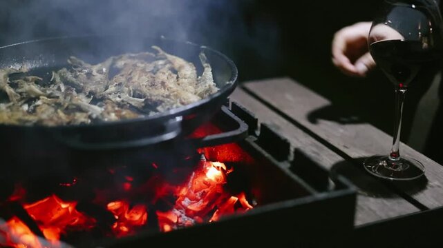 Nighttime slow-motion grilling scene with fish cooking on a barbecue grill, glass of wine reflecting warm firelight and a partially visible person preparing food outdoors for a cozy evening atmosphere