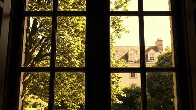 View through a dark wooden mullioned window to a sunny garden with lush green tree foliage and a traditional house with dormer windows in the background