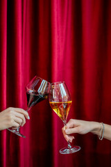 Two hands holding red and white wine in elegant glasses, toasting in a celebratory moment against a luxurious red curtain background