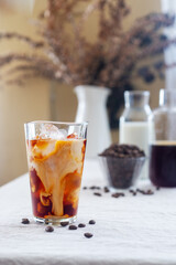 A glass of cold brew coffee with milk and ice sits on a table covered with a natural linen tablecloth.
