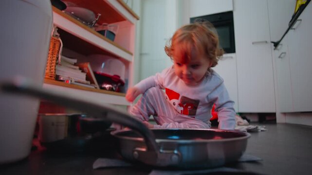 Toddler playing with kitchen cookware on floor reaching toward small pot with gentle curiosity while navigating space around shelves and utensils in simple indoor environment