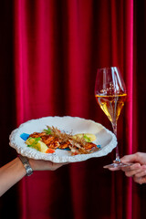 shallow depth of field of grilled salmon with garnishes on a white plate being held over a deep red curtain. A hand holds a glass of amber-colored sparkling wine next to the dish