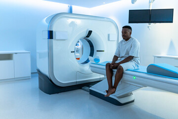 African man sitting on gantry table in exam room, in gown, blue pad visible, copy space