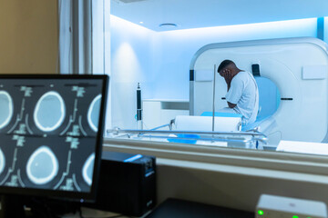 Patient sitting on CT table holding head, control-room monitor showing brain scans, blue gantry