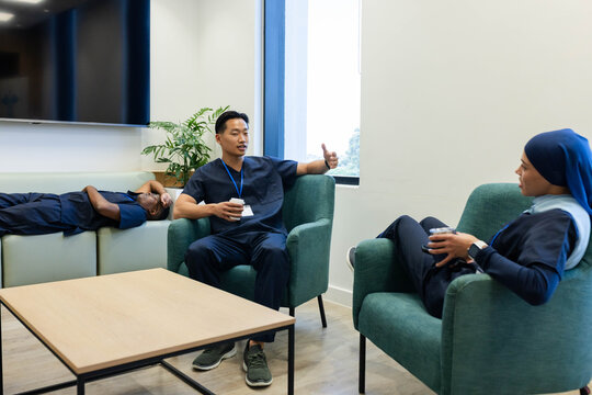 Diverse coworkers in navy scrubs taking break at staff lounge, holding coffee cups with ID lanyards