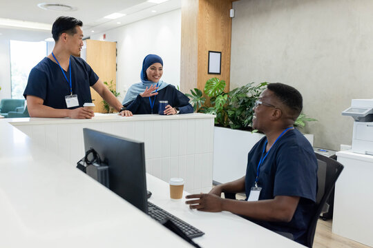 Diverse coworkers wearing navy scrubs talking at clinic reception desk with coffee cups, copy space