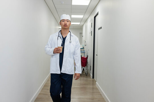 Asian male doctor walking hospital hall navy scrubs, white lab coat, holding paper cup, stethoscope