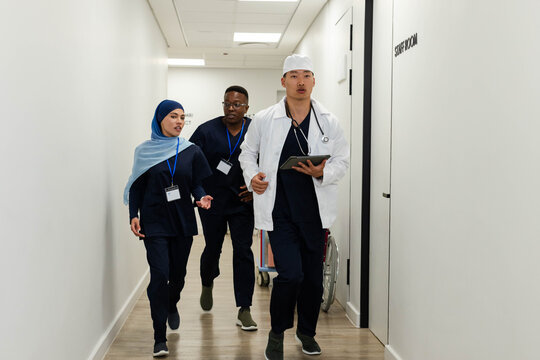 Diverse medical staff walking hospital corridor, Asian physician holding tablet, wearing white coat