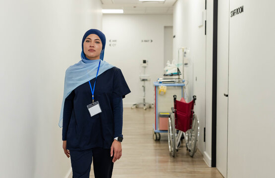 Female nurse standing in hospital corridor wearing navy scrubs, hijab, ID badge, wheelchair nearby