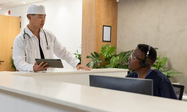 Diverse male coworkers discussing patient checkin at clinic reception desk with lab coat, tablet