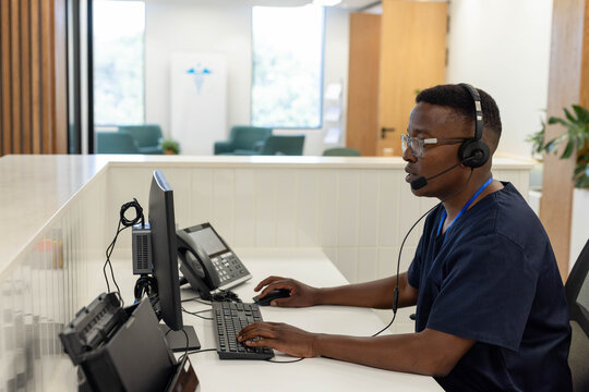 Man in navy scrubs sitting at reception desk typing on wired keyboard with headset, copy space