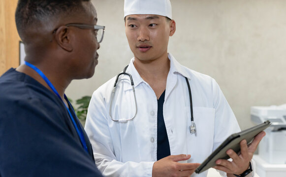 Diverse male staff discussing case in clinic, doctor in white coat with stethoscope holding tablet