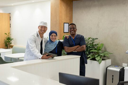 Diverse medical team in scrubs and coat standing at clinic reception holding tablet and stethoscope