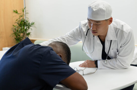 Asian male doctor in coat leaning, placing hand on male African patient at round table, stethoscope