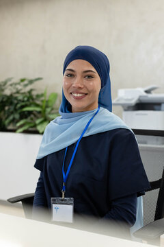 African American woman sitting at clinic reception desk, smiling, wearing navy hijab and badge
