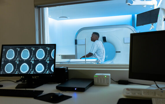 African American man in gown sitting on MRI table in control room, monitors showing brain scans