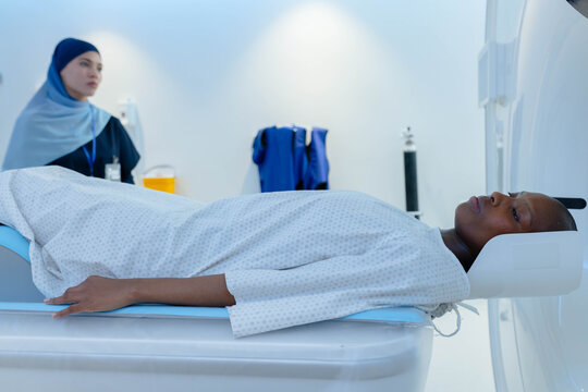 African female patient lying on exam table in CT scanner, female tech in scrubs monitoring