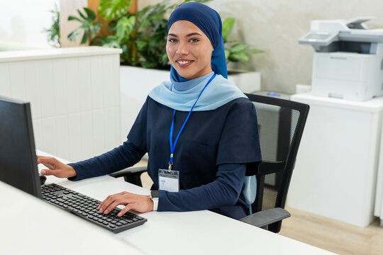 Adult female in navy scrubs light blue hijab typing at clinic reception with keyboard and lanyard