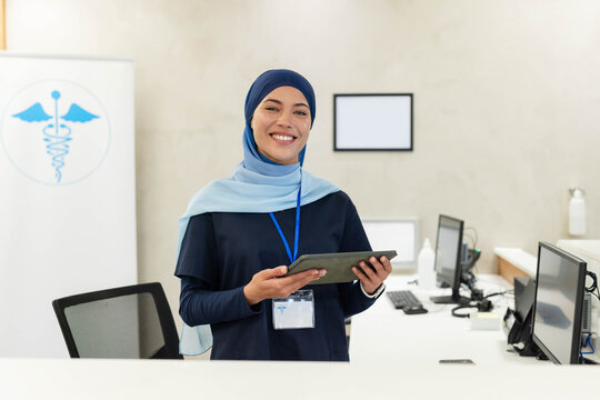 Female clinic worker wearing navy scrubs, blue hijab, holding tablet and badge at clinic reception