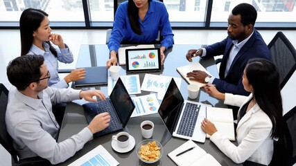 Diverse business team meeting around a table analyzing charts and graphs on laptop and tablet during a corporate strategy session - Powered by Adobe