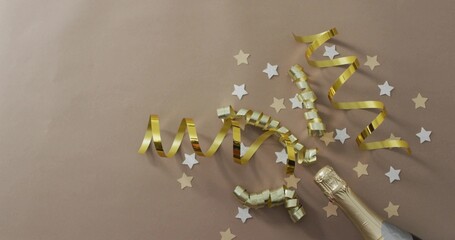 Arranging gold ribbons and star confetti near champagne bottle neck on kraft paper, copy space