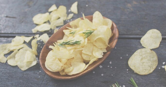 Fototapeta Displaying wooden oval bowl holding potato chips on dark tabletop, with rosemary and coarse salt