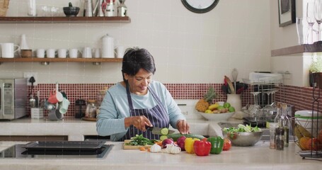Chopping senior Indian cook in striped apron slicing cucumber at kitchen island, with cutting board © vectorfusionart