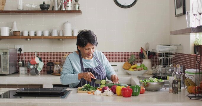 Chopping senior Indian cook in striped apron slicing cucumber at kitchen island, with cutting board