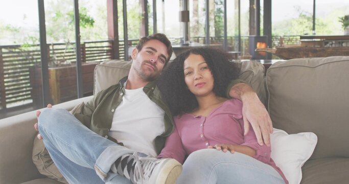 Sitting couple wearing casual clothes cuddling on light-beige sofa in home lounge, with glass doors