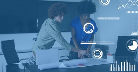 Leaning light-shirted man and blue-bloused woman pointing at blueprint in meeting room, with laptop