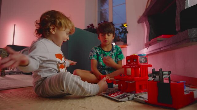 Young children playing together on floor with toy fire station playset during quiet indoor moment, older brother watching toddler concentrate while building small pieces with focused hands