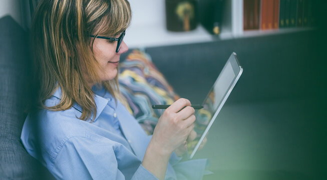 Woman working remotely. Uses stylus on tablet on sofa. Comfortable work from home. Relaxed workspace - Powered by Adobe