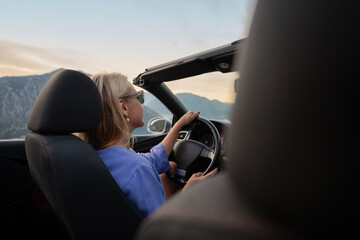 Young woman driving convertible along scenic ocean coast and mountain road at sunset, carefree summer vacation, travel freedom and adventure on coastal highway