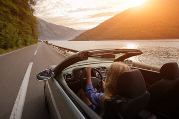 Young woman driving convertible along scenic ocean coast and mountain road at sunset, carefree summer vacation, travel freedom and adventure on coastal highway