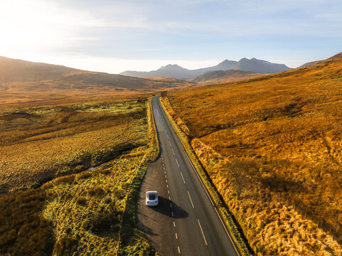 Aerial view of a lone car navigates the stark, black ribbon of road cutting through golden fields towards the distant peaks of Snowdonia, Snowdon, Wales, United Kingdom.