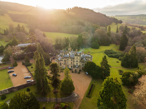 Aerial view of a grand, ornate building basking in the golden glow of the sunset, nestled amidst lush green fields and shadowed trees, Bala, Wales, United Kingdom.