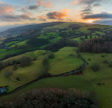 Aerial view of a winding road cutting through vibrant green hills, under a sky painted with sunset hues over Bala, Wales, United Kingdom.