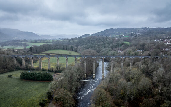 Aerial view of a grey stone aqueduct soaring over the flowing River Dee, a stark contrast to the muted greens and browns of the surrounding landscape, Llangollen, Wales, United Kingdom.