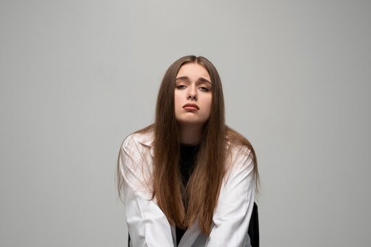 Sad tired woman in white coat looking down with bored facial expression on neutral gray studio background